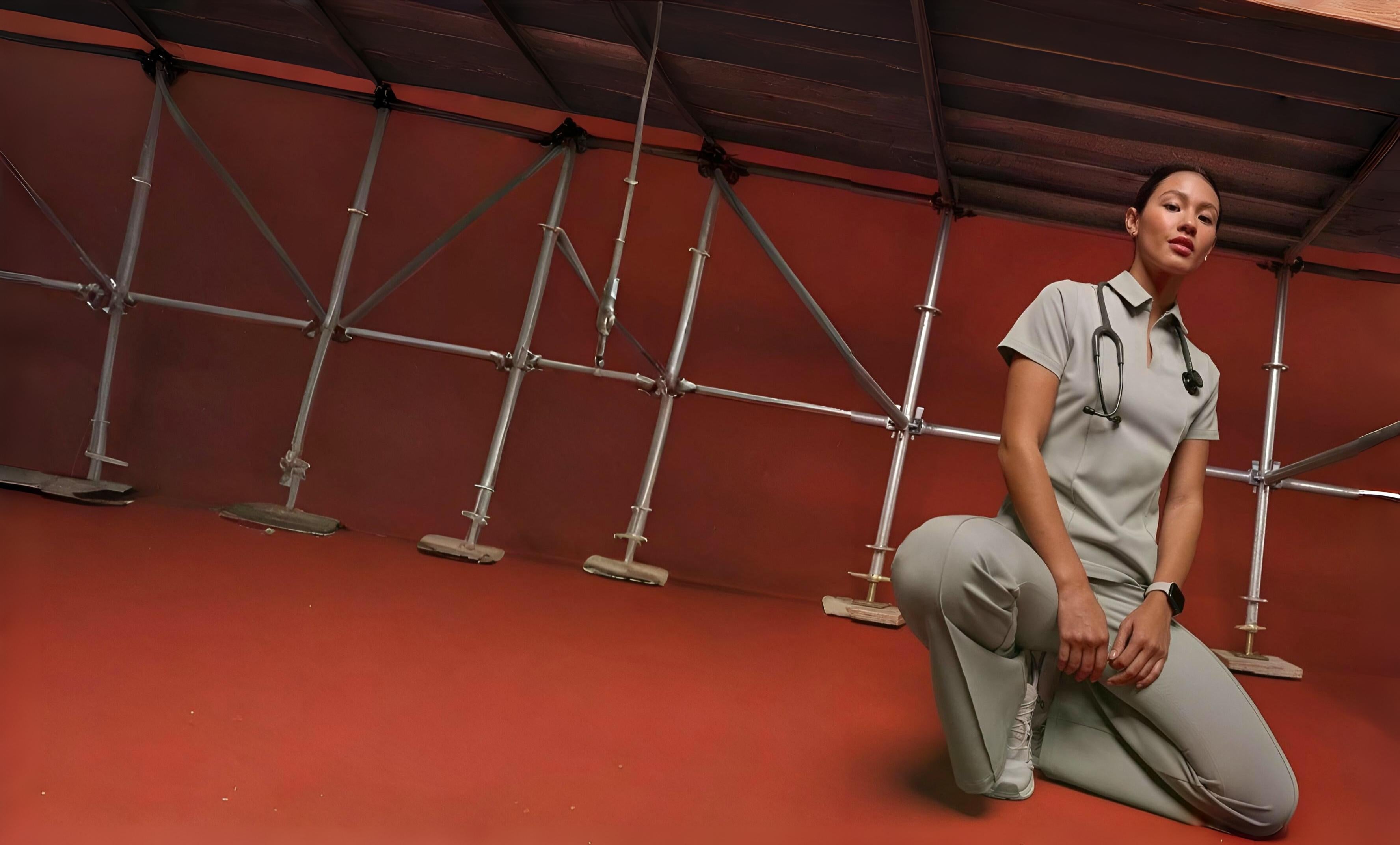 Person in medical attire kneeling on a red floor with metal beams in the background